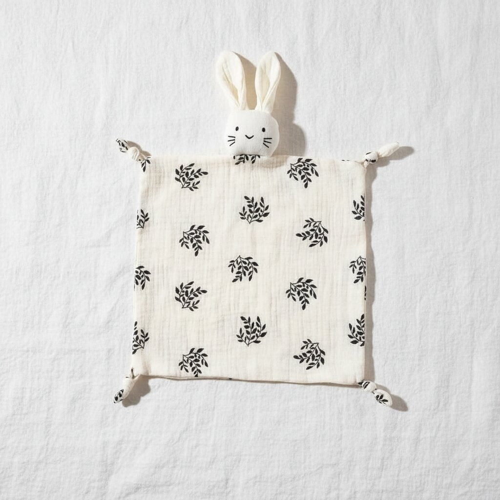 Doudou lapin en gaze de coton motif feuilles botaniques pose a plat sur un drap de lit blanc